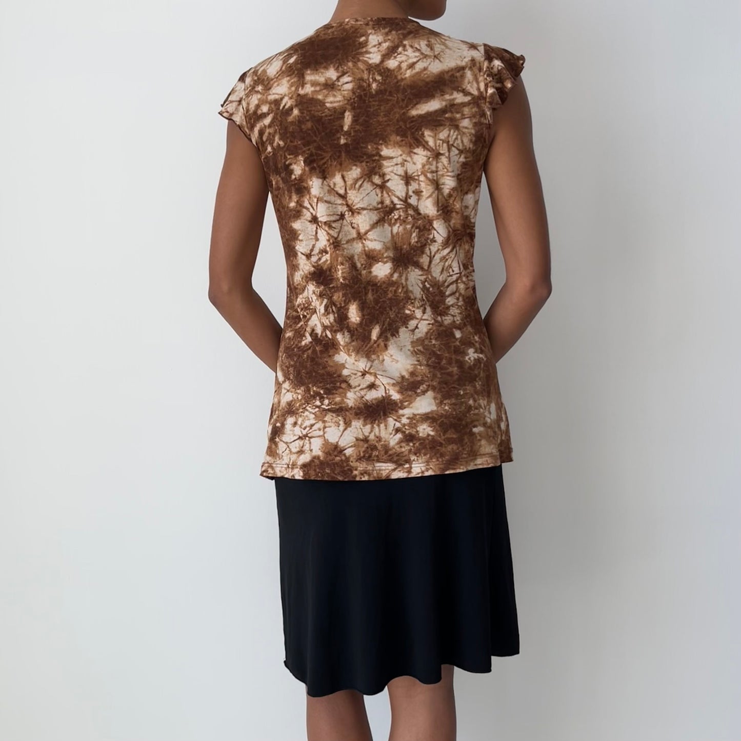 A woman shown from the back wearing a brown and beige tie-dye short-sleeve top and a black skirt, standing against a plain light gray background.