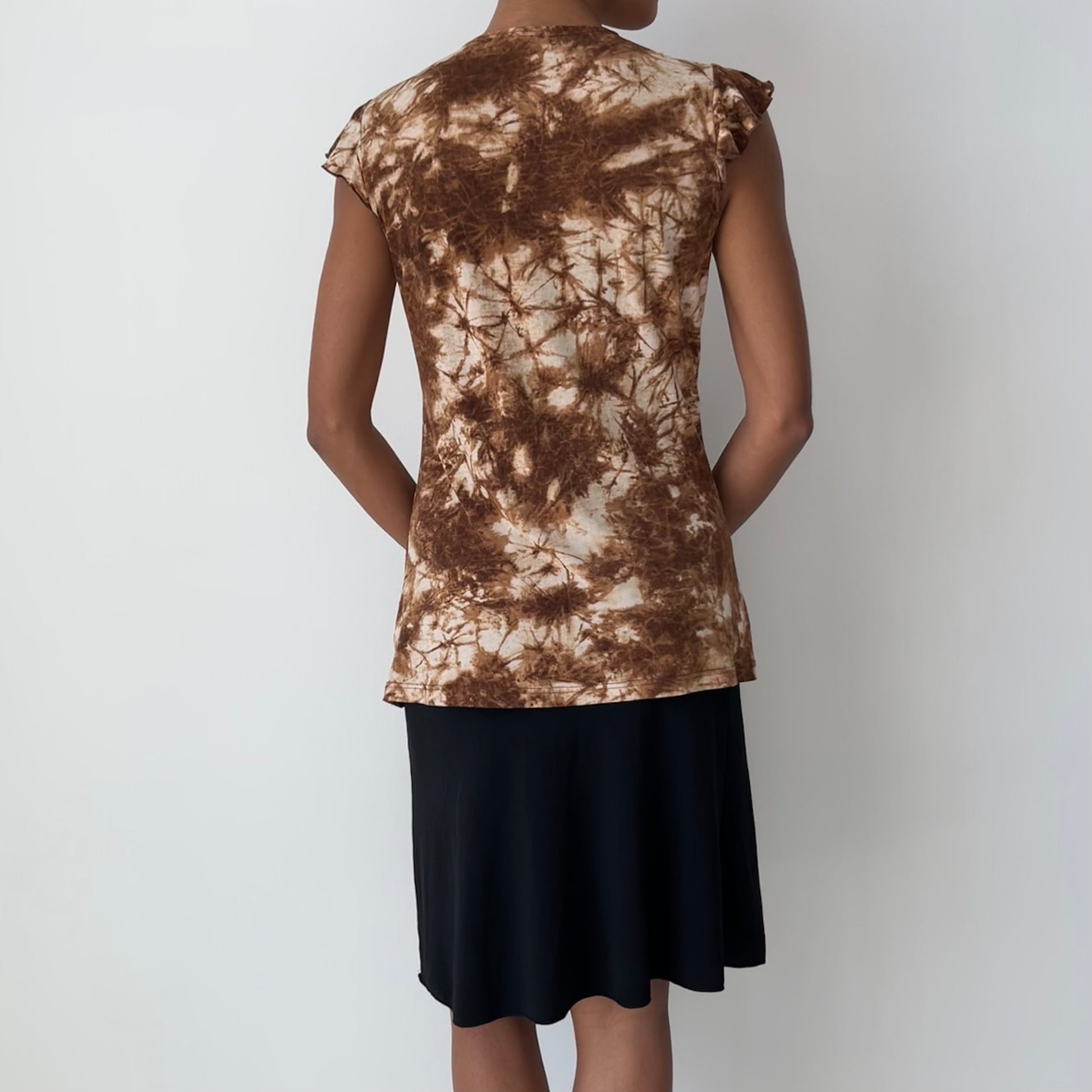 A woman shown from the back wearing a brown and beige tie-dye short-sleeve top and a black skirt, standing against a plain light gray background.